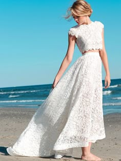 a woman is standing on the beach in a white lace dress and matching heels with her hair blowing in the wind