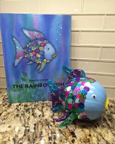 the rainbow fish is next to a book and some decorations on a counter top in front of a tile wall