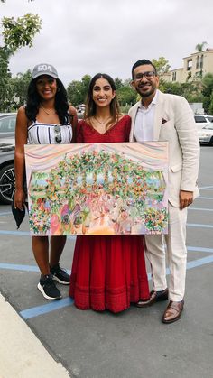 three people standing next to each other holding a large painting in the middle of a parking lot