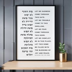 a white framed poster with the words in hebrew on it next to a potted plant