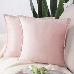 two pink pillows sitting on top of a white couch next to a book and potted plant