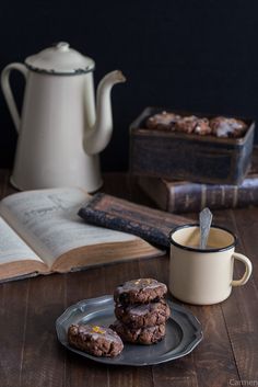 three cookies on a plate next to an open book and a tea pot with a spoon in it