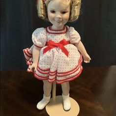 a doll with blonde hair wearing a white dress and red bow on her head is standing on a wooden table