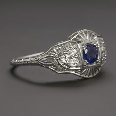 This beautiful and unique ring features a richl blue sapphire set in a diamond studded vintage style design that will never go out of style! The sapphire has a richly saturated blue hue that contrasts strikingly with the 14k white gold setting. Utterly classic and romantic, the antique style setting is richly textured with intricate filigree and milgrain. The effect is chic but also dreamy and softly feminine, a perfect choice for a timelessly stylish right hand ring or alternative engagement ri Diamond And Blue Sapphire Wedding Ring, Blue Platinum Art Deco Diamond Ring, Classic Blue Diamond Ring, Blue Single Cut Diamond Jewelry In Platinum, Platinum Jewelry With Blue Single Cut Diamonds, Blue Platinum Jewelry With Single Cut Diamonds, Art Deco Blue Diamond Ring, Blue Diamond Art Deco Ring, Blue Art Deco Diamond Ring