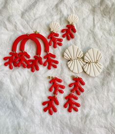 three red and white earrings are laying on a table cloth next to two seashells