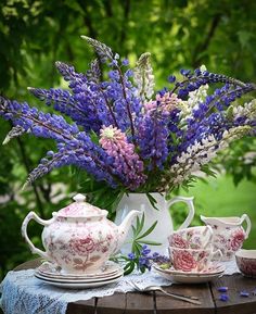 a table topped with two tea cups and a vase filled with purple flowers on top of a wooden table