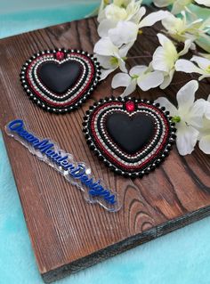 two black and red heart shaped earrings sitting on top of a wooden board next to white flowers