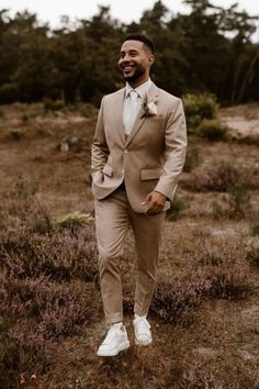 a man wearing a tan suit and white shoes standing in a field with purple flowers