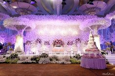 an elaborately decorated wedding stage with purple flowers and greenery on the ceiling, along with two tiered cake stands