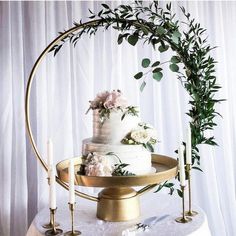 a three tiered wedding cake with greenery and candles on a gold stand in front of a white curtain