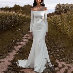 a woman in a white dress standing on a dirt path with grass and bushes behind her