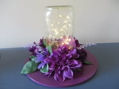 a glass jar with lights and flowers in it on a purple plate next to a wall