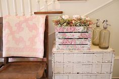 an old dresser with flowers and a quilt on it next to a wooden chair in front of a stair case