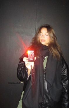 a woman taking a selfie with her camera in front of a black wall and wearing a leather jacket