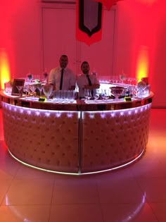 two men are standing behind a bar with wine glasses on it and red lights in the background