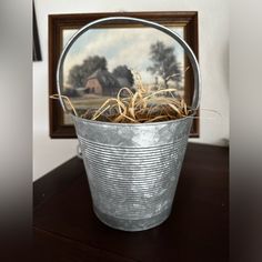 a metal bucket filled with grass sitting on top of a wooden table next to a painting