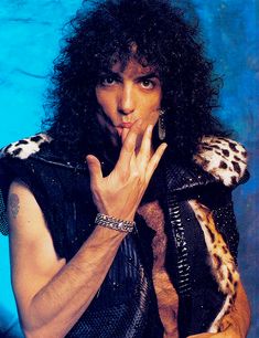 a man with long black hair wearing a leopard print vest and holding his hands up to his mouth