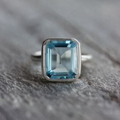 A handmade Sky Blue Topaz Emerald Cut Ring in Recycled Silver on a wooden surface. Modern Octagon Topaz Gemstone Ring, Modern Octagon Topaz Ring, Timeless Blue Topaz Ring Perfect As Gift, Timeless Blue Topaz Ring As Gift, Timeless Blue Topaz Ring For Gift, Modern Rings, Asscher Engagement Ring, Asscher Cut Engagement Rings, Emerald Cut Ring