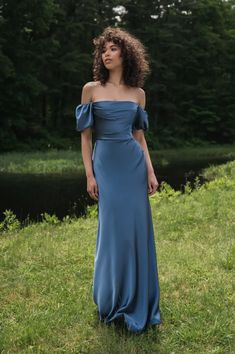 a woman wearing a blue dress standing in the grass near a river and trees, looking off to the side