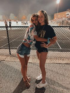 two young women standing next to each other on a tennis court at night with people in the background