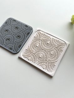 two square coasters sitting on top of a table next to a vase with flowers