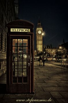 a telephone booth sitting on the side of a street next to a clock tower at night