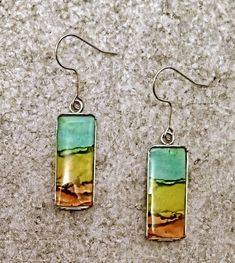 two square shaped earrings on top of a gray surface, one with a green and yellow rectangle in the middle