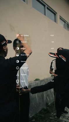 two men in black hoodies are standing near a building and pointing at something on the wall