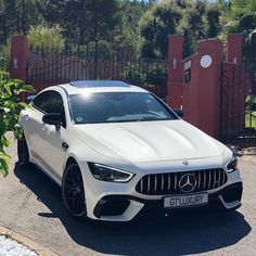 a white mercedes benz coupe parked in front of a gated driveway with trees and bushes