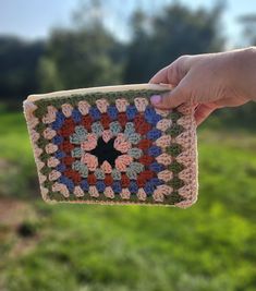 a hand holding up a crocheted square in front of some grass and trees