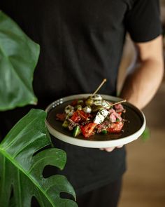 a man holding a plate with meat and vegetables on it in front of a plant