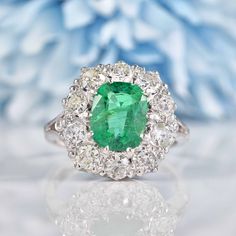 an emerald and diamond ring on a white surface with blue flowers in the back ground