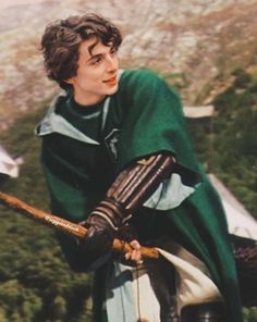 a young man dressed in medieval clothing holding a wooden stick and looking off into the distance