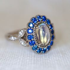 Mesmerizing Blue Moonstone Halo Ring. Moonstone engagement ring. Blue sapphires & diamonds. Edwardian Moonstone. 14K White Gold. US 7.25 A beautiful and unique moonstone ring, created with precision cut sapphires in 14K white gold, a luxurious and high-quality piece of jewelry, perfect to express your love. 💎 PREMIUM RING -- A brand new moonstone ring, personally designed and made with the highest quality of 14K white gold.  ✨ CLASSIC BRILLIANCE -- Designed with a beautiful blue moonstone at th Luxury Vintage Moonstone Ring As A Gift, Luxury Victorian Moonstone Ring Gift, Art Deco Multi-stone Oval Sapphire Ring, Art Deco Oval Multi-stone Sapphire Ring, Heirloom Oval Moonstone Ring, Vintage Multi-stone Oval Sapphire Ring, Vintage Oval Multi-stone Sapphire Ring, Celestial Multi-stone Oval Moonstone Ring, Blue Oval Multi-stone Cluster Ring