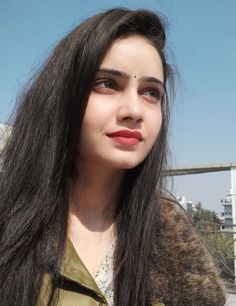 a woman with long black hair wearing a brown jacket and looking at the camera while standing in front of a bridge