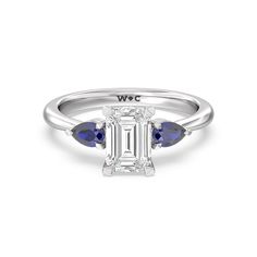 a white gold ring with two blue sapphire stones on the band and an emerald cut diamond