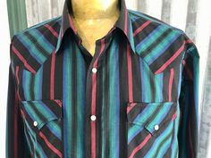 A classic cowboy style shirt in a bold colour palette. This vintage 1980's long sleeve shirt has traditional western details, such as pearl snap closures, peaked front and back yokes and two front pockets with peaked flaps. Features a striped design in deep red, emerald green, blue and black. Size Large. Measurements: Chest: 124 cm / 49 inches Length: 78 cm / 31 inches                                                                                  Shoulders: 48 cm / 19 inches                                                                                       Sleeve: 63 cm / 25 inches This item is one of a kind. BUYING VINTAGE: While all care is taken to state any obvious faults, please note that when you buy a vintage or preloved item, they may show small signs of ageing, wear and/or re Fitted Long Sleeve Shirt For Western-themed Events, Black Long Sleeve Western Shirt, Western Style Black Button-up Shirt, Western Black Button-up Shirt, Black Western Button-up Shirt, Western Long Sleeve Tops With Snap Buttons, Western Style Black Shirt For Ranch, Western Style Shirt For Summer Rodeo, Western Black Shirt For Ranch