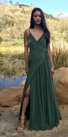 a woman in a green dress standing next to some rocks and water with her legs crossed