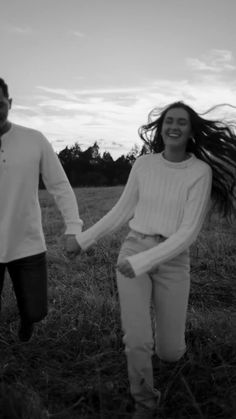 a man and woman holding hands walking through a field with long hair in the wind