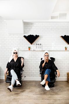 two women sitting on chairs in front of a white tiled wall with shelves and shelving