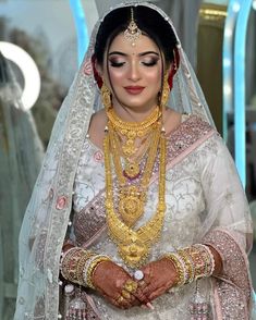 a woman in a white and gold bridal outfit with jewelry on her neck, standing next to a mirror