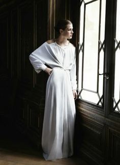 a woman standing in front of a window wearing a white dress