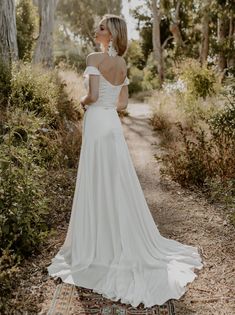 a woman in a white wedding dress standing on a path with trees and bushes behind her