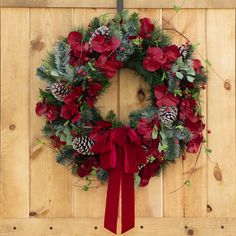 a christmas wreath with red flowers and pine cones hanging on a wooden wall, surrounded by greenery