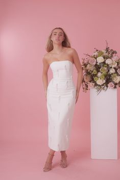 a woman standing next to a tall white vase with flowers in it on a pink background