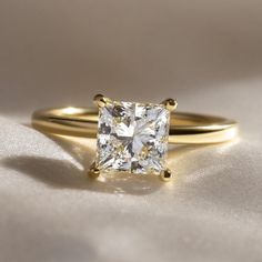 a gold ring with a princess cut diamond in the center on a white cloth background