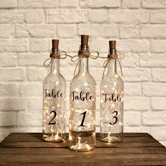 three glass bottles with lights in them sitting on a wooden table next to a brick wall