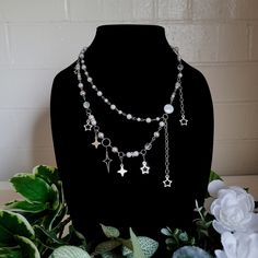 Introducing our Celestial Dreams Necklace - a whimsical, multi-layered piece that captures the enchantment of the night sky. This necklace features two distinct chains, each adorned with clear, iridescent beads that shimmer like distant stars. The upper chain is a delicate strand, with small beads evenly spaced for a subtle sparkle. The lower chain, in contrast, is a statement piece. Larger beads are interspersed with a variety of star and moon charms, each one unique in size and design. Two shooting star charms add a dynamic touch, their tails trailing behind them as if caught in mid-flight. This necklace is perfect for those who find beauty in the cosmos, or anyone who wants to add a touch of magic to their everyday look. Whether you're dressing up for a special occasion or simply want t Silver Star-shaped Charm Necklaces For Party, Silver Star Charm Necklaces For Party, Whimsical Silver Dangle Charm Necklaces, Whimsical Silver Necklaces For Party, Whimsical Silver Necklace For Party, Celestial Dangle Necklace With Star Charm, White Celestial Necklace With Star Charm, Celestial Star-shaped Necklace For Party, Celestial Star Necklace For Party