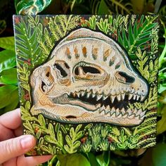 a hand holding up a painting of a dinosaur in the jungle with leaves and plants