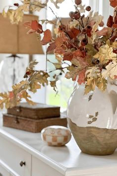 a white vase filled with lots of leaves on top of a table next to a lamp